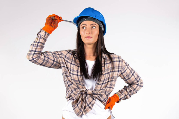 Female mechanic wearing blue hard hatholds a wrench and looks thoughtfully to the side. White background. High quality photo - 写真・画像