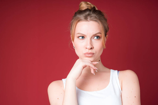 Beautiful blond woman with bun looks disapprovingly or accusingly in white tank top isolated on red background. - Foto, imagen