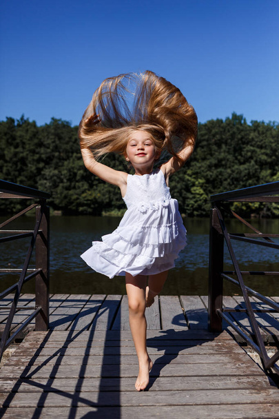 A six year old girl is wearing a white dress. Beautiful long hair. Walk near the river on the background of the forest - Photo, Image
