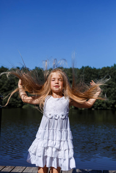A six year old girl is wearing a white dress. Beautiful long hair. Walk near the river on the background of the forest - Photo, Image