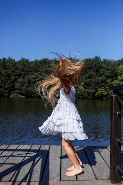 Egy hat éves lány fehér ruhát visel. Gyönyörű hosszú haj. Séta a folyó mellett az erdő hátterében - Fotó, kép