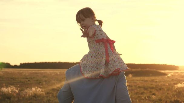 The little daughter rides on neck of her beloved daddy in sun. Happy family dad and child play on the field in rays of a warm sunset. Father and child travel in park in summer. Healthy childhood. - Photo, Image