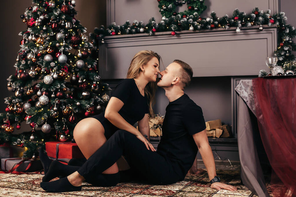 Sexy young woman in black bodysuit and handsome man are sitting on the floor and hugging. Young beautiful couple celebrating Christmas together at home near the Christmas tree. New Year - Photo, Image