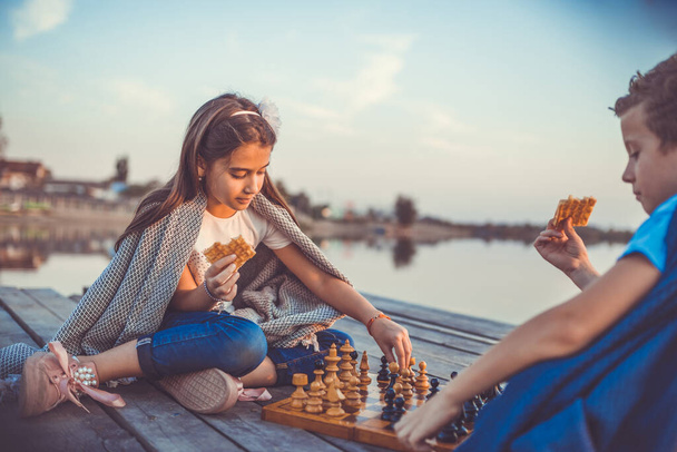 Dos amiguitos, niño y niña divirtiéndose mientras juegan al ajedrez sentados junto al lago por la noche. Los niños están jugando. Amistad. Enfoque selectivo. - Foto, imagen