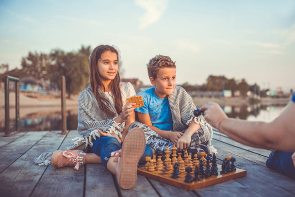 Δύο νεαροί χαριτωμένοι μικροί φίλοι, αγόρι και κορίτσι διασκεδάζουν παίζοντας σκάκι καθισμένοι δίπλα στη λίμνη το βράδυ. Παίζουν τα παιδιά. Φιλία. Επίλεκτη εστίαση. - Φωτογραφία, εικόνα