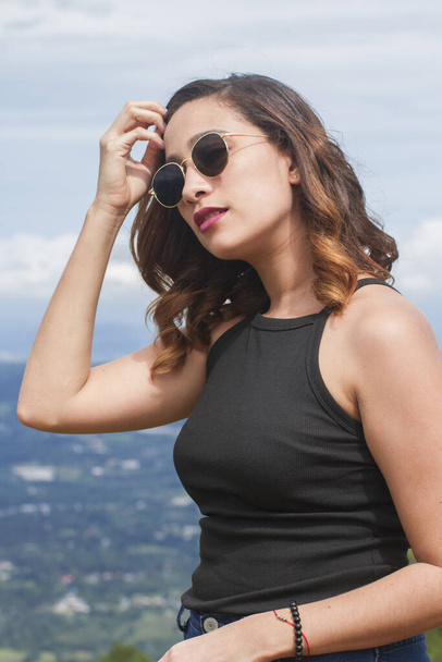 half body shot of one attractive hispanic young woman or girl with sunglasses caressing her hair outdoor in a sunny summer day with nature in background - Photo, Image