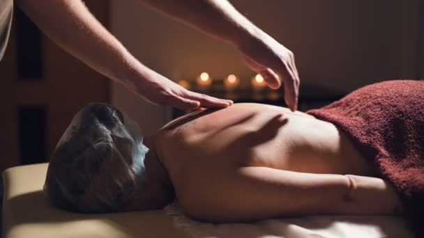Young male massage therapist is doing finger massage of a woman back with a tattoo in a massage room with dim light on the background of candles. Low key premium massage concept - Video