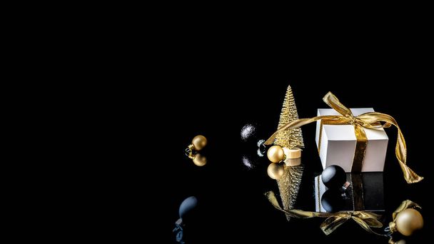 Happy Christmas. White gift with golden bow, gold balls and new year tree in xmas decoration on dark background for greeting card. Christmas, winter, new year concept - Photo, Image