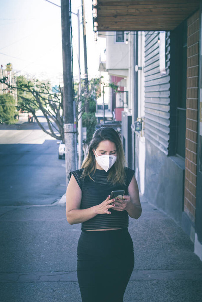 элегантная латиноамериканка в маске для лица с помощью мобильного телефона на улице - Фото, изображение