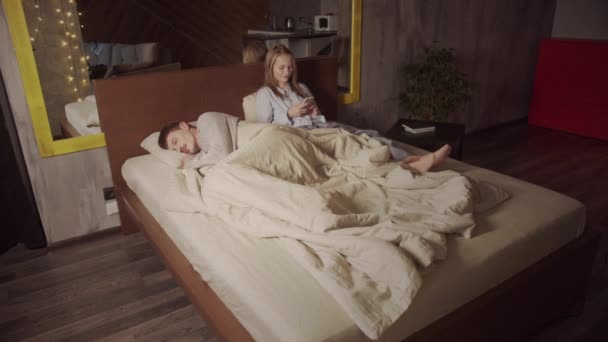 Una giovane bella ragazza è sdraiata sul letto in pigiama con il marito addormentato. Chiacchierare e scorrere attraverso il feed del social network instagram. - Filmati, video