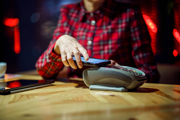 NFC terminali yakınlarında telefonu olan olgun bir kadın kahve tezgahında cep telefonu ödemesi yapıyor. Üst düzey kadın müşteriler de kafeteryada cep telefonuyla ödeme yapıyor. POS uçbirimi. - Fotoğraf, Görsel