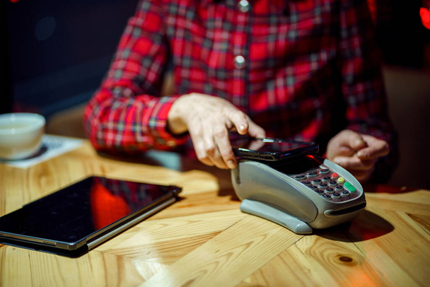 Női érett ügyfél kezében telefon közelében NFC terminál, hogy érintésmentes mobil fizetés a kávézó pult, idősebb nő ügyfél fizet a kávézóban mobiltelefonnal keresztül pos gép. POS terminál. - Fotó, kép