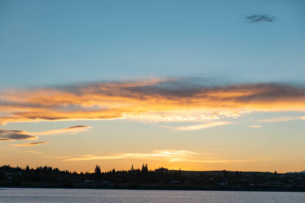 太陽が焦点を合わせる時に、レンズ状の雲は、シルエットの前景の上方とテカポ湖の上の澄んだ青い空の明るいオレンジ色になります。. - 写真・画像