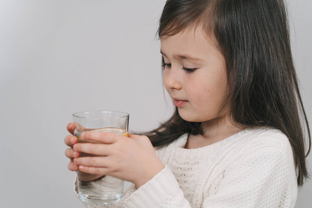 Το παιδί πίνει νερό από ένα ποτήρι. Ένα κορίτσι με σκούρα μαλλιά κρατάει ένα ποτήρι νερό.. - Φωτογραφία, εικόνα