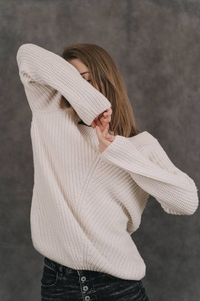 A girl in a light sweater on a gray background. Womens hands close-up. The girl covers her eyes with her hands - Фото, изображение