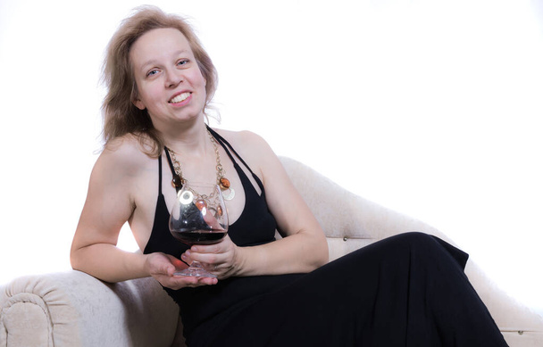 Cheerful 40 years old woman in black dress having relax on sofa with glass of wine - Фото, зображення