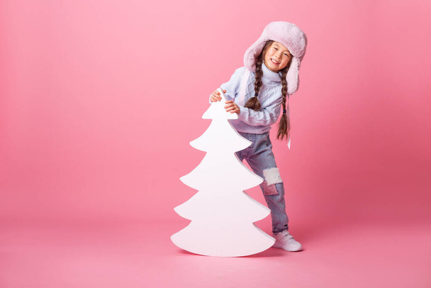 asian girl with fir tree shaped decoration wearing big furry hat posing at pink studio - Foto, afbeelding