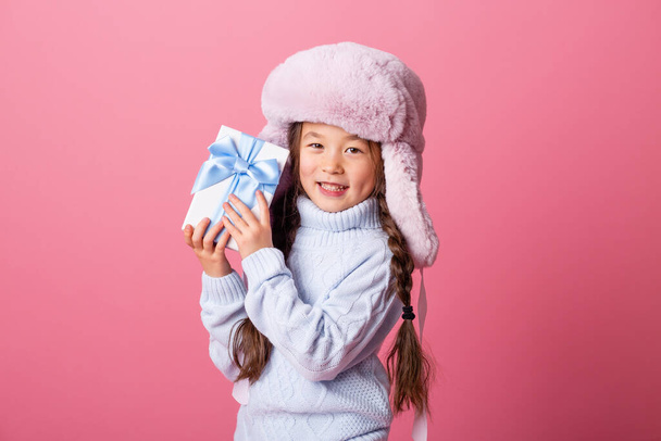 pequeña chica asiática en invierno sombrero posando con caja de regalo sobre fondo rosa  - Foto, Imagen