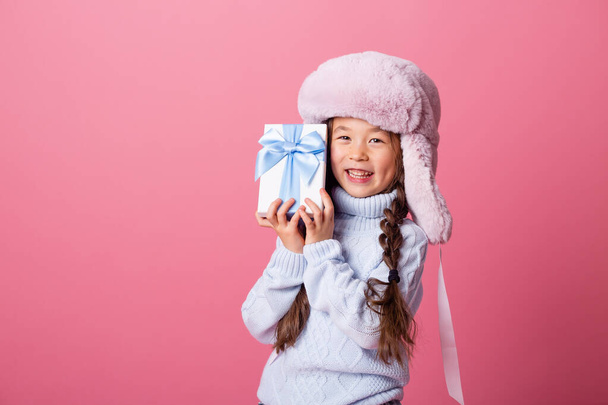 petite fille asiatique en chapeau d'hiver posant avec boîte cadeau sur fond rose  - Photo, image