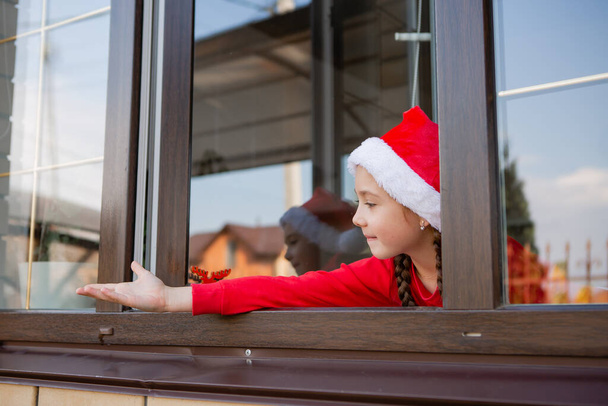 κορίτσι με το καπέλο santa κοιτάζοντας έξω από το παράθυρο και φθάνοντας το χέρι - Φωτογραφία, εικόνα