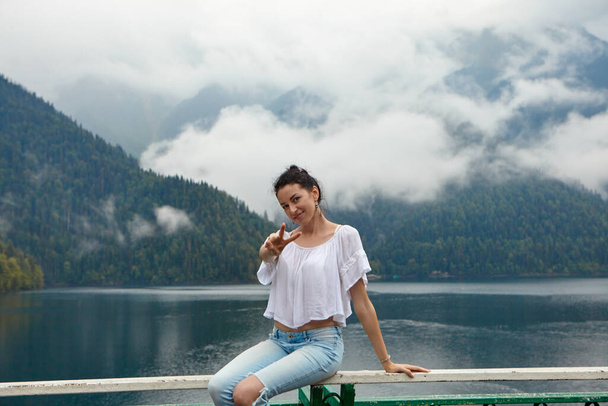 Ένα κορίτσι με λευκά ρούχα σε φόντο βουνίσιας λίμνης με μπλε νερά και βουνά με σύννεφα. Ιστορικό. - Φωτογραφία, εικόνα