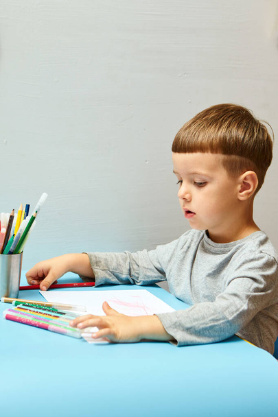 Söpö pieni poika vaaleat hiukset kiinnittää värillisiä kyniä kotona. Piirtää sinisessä pöydässä. - Valokuva, kuva