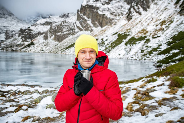 Hombre viajero en una chaqueta roja y gorra amarilla sostiene una taza de té caliente de metal en un hermoso paisaje de montaña. - Foto, Imagen