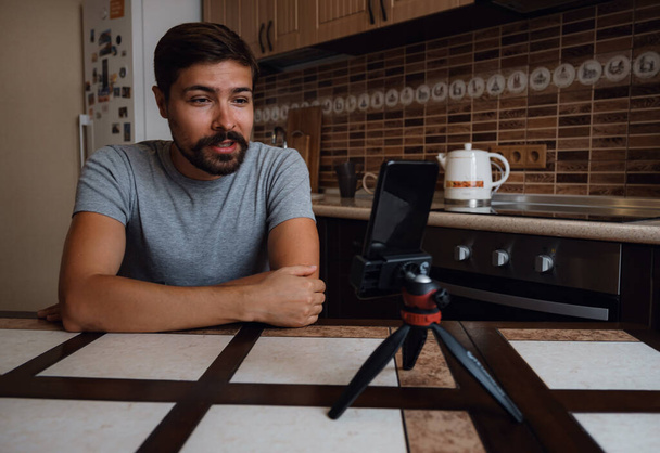 自宅のスマートフォンでビデオ通話をしている若い男。バーチャルハウスパーティーだ。自信満々の若い男vloggerインフルエンサーは自宅でソーシャルメディアビデオブログトークを撮影 - 写真・画像