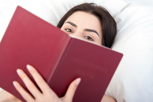 Закрыть молодую женщину, читающую в постели
 - Фото, изображение