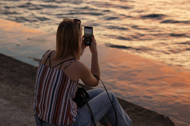 Chica fotografías turísticas en el teléfono un hermoso paisaje acuático al atardecer. - Foto, Imagen