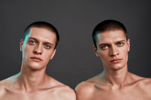 Portrait of young half naked twin brothers looking at camera while standing isolated over dark grey background, front view - Foto, Imagem