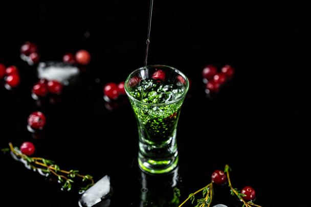shot αψέντι χύνεται σε ένα ποτήρι. μπουκάλι αψέντι με cranberries και lime που απομονώνονται σε μαύρο φόντο. χώρος για κείμενο. - Φωτογραφία, εικόνα