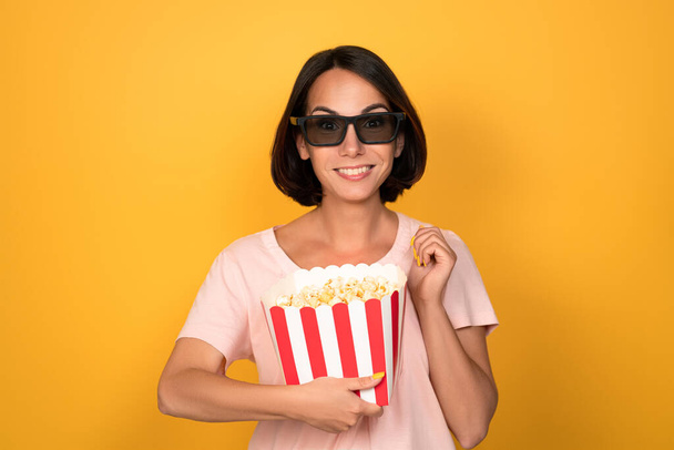 Bucket of popcorn and stereo 3D glasses in hands of a young brunette woman. Cinema concept. Isolated on yellow background - Foto, Imagen