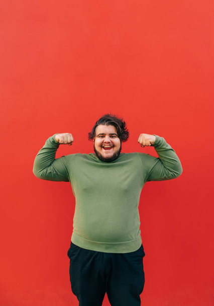 Vertical shot of a strong young fat hispanic man demonstrating his power by raising his hands and clenching his fists, showing muscles, smiling and standing on a red background. - Foto, Bild