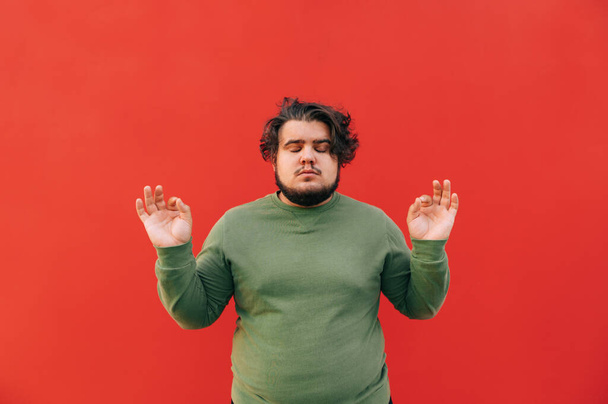 Ένας νεαρός Ισπανός με πράσινη μπλούζα διαλογίζεται και συγκεντρώνεται, στέκεται πάνω σε κόκκινο φόντο με τα μάτια κλειστά, υψώνοντας τα χέρια του σε μια καλή χειρονομία.. - Φωτογραφία, εικόνα