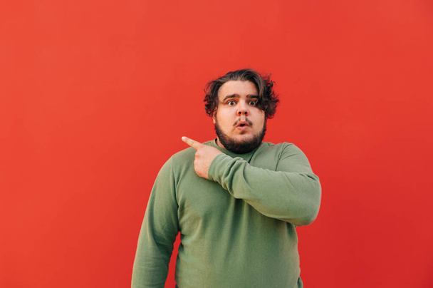 Удивленный толстый латиноамериканец в зеленой толстовке показывает пальцем назад на красном фоне, выглядя шокированным и изумленным. - Фото, изображение