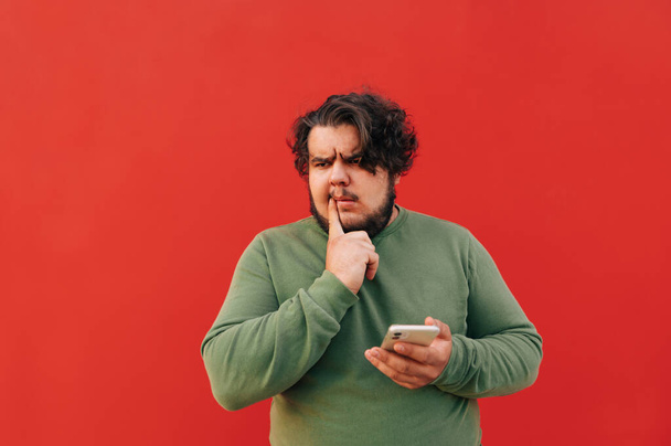 Huomaavainen latino kaveri matkapuhelimella ilmaisee ajattelevaa elettä, katsoen sivuun, rypistäen ja hämmentyneenä älypuhelinta käyttäessään. - Valokuva, kuva