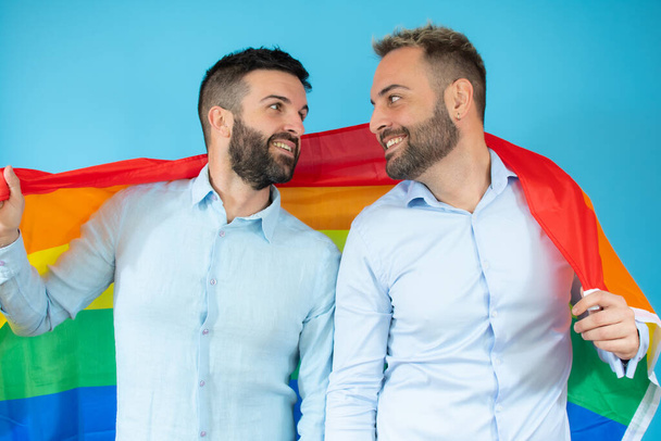 A young gay couple holding a flag over blue background. - Φωτογραφία, εικόνα