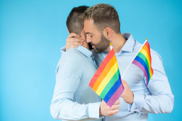 Ein junges schwules Paar hält eine Fahne und umarmt sich vor blauem Hintergrund. - Foto, Bild