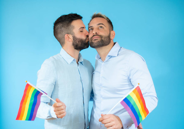 Ένα νεαρό γκέι ζευγάρι που κρατάει μια σημαία πάνω από μπλε φόντο.. - Φωτογραφία, εικόνα