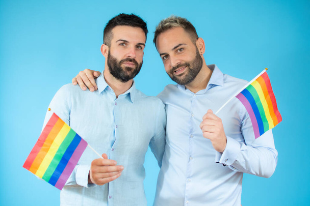 若いゲイのカップルが青い背景に旗を掲げて. - 写真・画像
