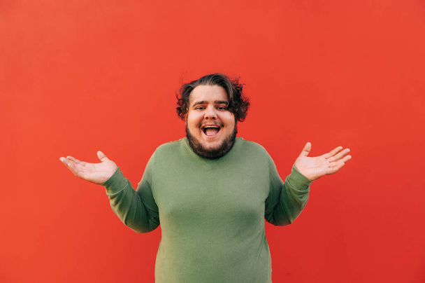 Cheerful and funny fat young hispanic man is expressing a confusion by a gesture, throwing up his hands, smiling, standing on an isolated red background. - Photo, Image