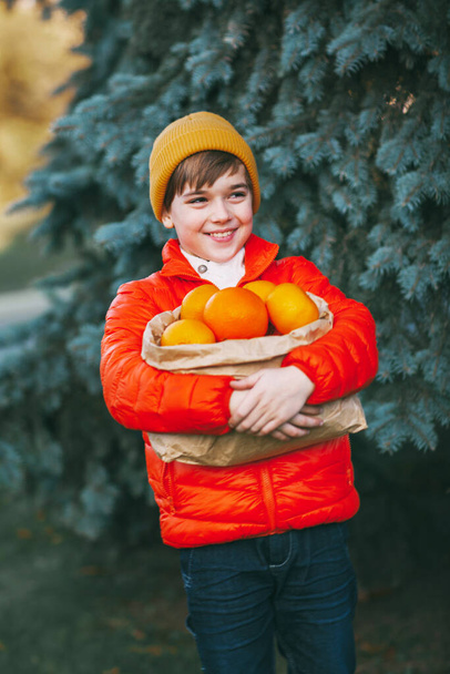 Un ragazzo con una giacca arancione brillante e un cappello giallo tiene in mano un grande pacchetto con arance sullo sfondo di un albero di Natale con una faccia sorpresa. Prepararsi per Natale, Capodanno - Foto, immagini