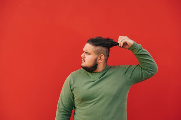 Widok z boku portret przystojnego mężczyzny z nadwagą w zielonej bluzie na czerwonym tle rozwiązującym włosy, patrzącego poważnie na bok. - Zdjęcie, obraz