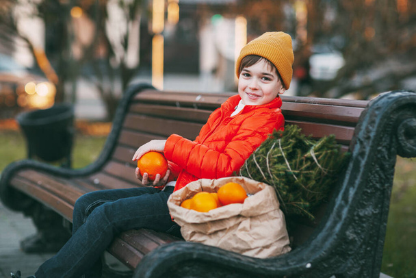 мальчик в ярко-оранжевой куртке и модной желтой шляпе сидит на скамейке, смеется, бросает апельсины в воздух, а рядом с ним маленькое дерево и большой мешок апельсинов. покупка для - Фото, изображение