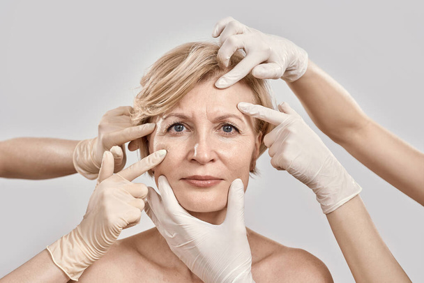 Beauty portrait of middle aged woman going under cosmetic treatment. Beautician hands in gloves checking female face skin isolated against grey background - Fotó, kép