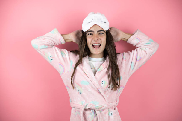 Jolie fille en pyjama et masque de sommeil sur fond rose relaxant et étirant, bras et mains derrière la tête et le cou souriant heureux - Photo, image