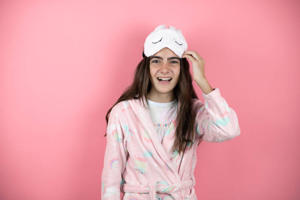 Όμορφο κορίτσι φοράει πιτζάμες και μάσκα ύπνου πάνω από ροζ φόντο σύγχυση και αναρωτιούνται για την ερώτηση. Αβέβαιο με αμφιβολία, σκέψη με το χέρι στο κεφάλι - Φωτογραφία, εικόνα