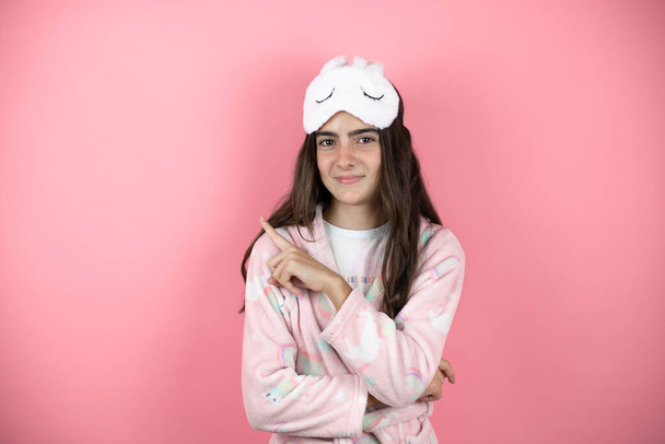 Chica bonita con pijama y máscara de dormir sobre fondo rosa sonriendo feliz señalando con la mano y el dedo a un lado - Foto, imagen