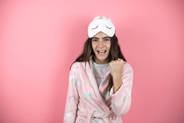 Chica bonita con pijama y máscara de dormir sobre fondo rosa enojado y loco levantando el puño frustrado y furioso mientras gritaba con ira. - Foto, Imagen